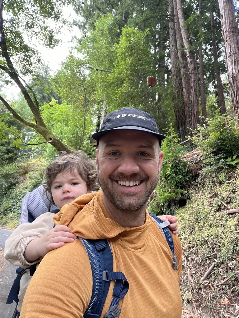 Clifton carrying a child in a hiking carrier beneath tall trees